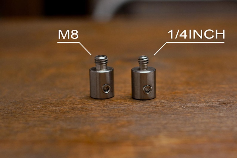 May include: Two small, cylindrical metal adapters with threaded tops. One is labeled "M8" and the other "1/4 INCH". The adapters have a small hole on the side. The background is a wooden surface.