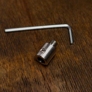 May include: A small, silver metal adapter with a threaded end and a hex key on a wooden surface. The adapter has a cylindrical shape with a hole on the side. The hex key is L-shaped.