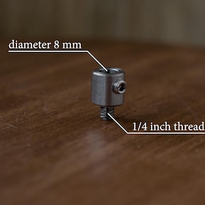 Puede incluir: Un conector de metal plateado con una rosca de 1/4 de pulgada y un di&aacute;metro de 8 mm. El conector est&aacute; sobre una superficie de madera.