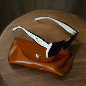 May include: Black sunglasses with white frames and a brown leather case. The sunglasses have the text "PRADA MILANO" printed on the frame.