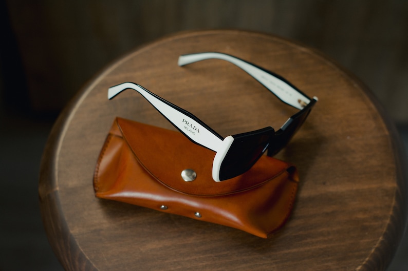 May include: Black sunglasses with white frames and a brown leather case. The sunglasses have the text "PRADA MILANO" printed on the frame.
