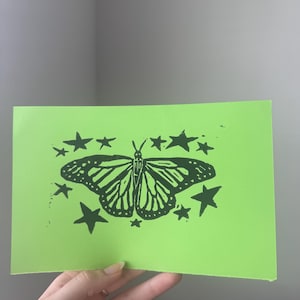 May include: A neon green card featuring a black butterfly design surrounded by stars. The butterfly has detailed wing patterns. The card is held in a person's hand.