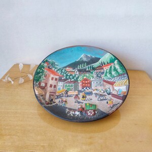 Peut inclure: Une assiette ronde en céramique peinte avec une scène de village dans les montagnes. L'assiette représente un ciel bleu, des arbres verts, des maisons brunes et des personnes en vêtements colorés.