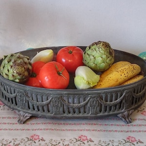 Op de afbeelding: Een donkergrijze metalen serveerschaal met sierlijke details, gevuld met verschillende soorten fruit en groenten, waaronder artisjokken, tomaten, bananen en sla.