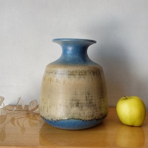 Peut inclure: Un vase en céramique bleu et beige avec une large ouverture et un corps arrondi. Le vase a un glaçage marbré et un aspect rustique.