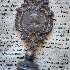 Op de afbeelding: Een vintage zilverkleurige metalen zegel met een gedetailleerd ontwerp. De zegel heeft een rond, versierd frame met een kleiner rond medaillon in het midden. Het medaillon toont het profiel van een vrouw met een kroon en de tekst "S. JOSEPH D'ARIMATEE". De zegel is bevestigd aan een ronde, zilverkleurige basis.