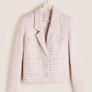 May include: A pale pink and white houndstooth blazer with a notched lapel, two front pockets, and three pearl buttons. The blazer is suspended on a wooden hanger. The brand name "GIACCA" is visible on the hanger.