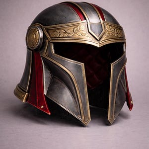 May include: A detailed, metallic helmet with a dark gray base, accented with gold trim and red stripes and accents. The helmet features a laurel wreath design and a circular emblem on the side, showcasing a blend of historical and futuristic aesthetics.