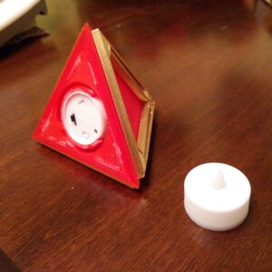 May include: A red triangular shaped object with a gold trim and a white circular button in the centre. A white round tea light is also shown on a wooden surface.