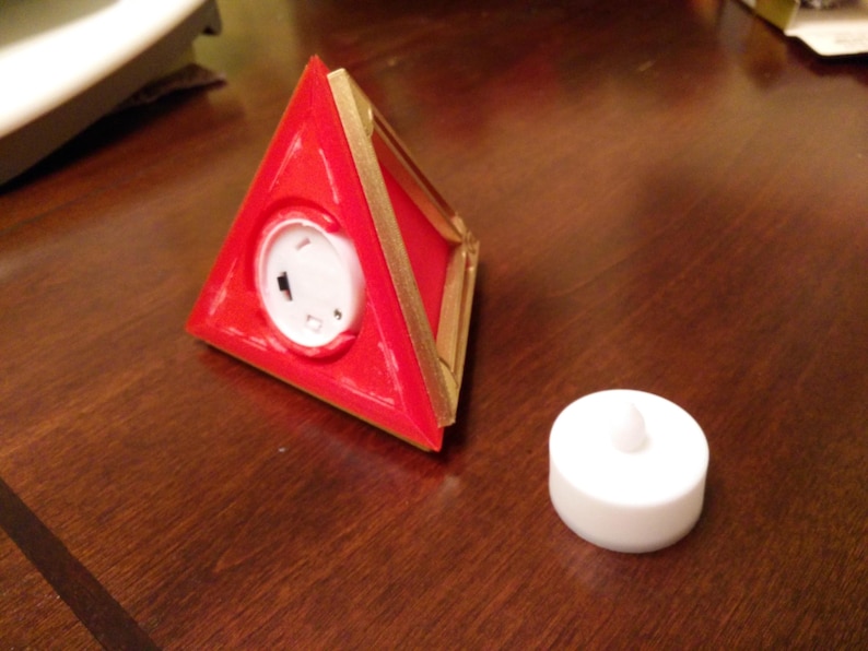 May include: A red triangular shaped object with a gold trim and a white circular button in the centre. A white round tea light is also shown on a wooden surface.