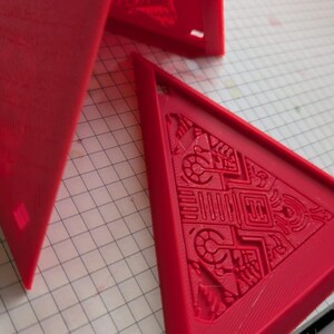 May include: Three red plastic triangular shapes with intricate designs on the surface. The shapes are arranged in a pyramid formation.