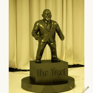 Può includere: Una statuetta premio dorata di una persona in abito in piedi su una base nera. La base ha il testo "Ihr Text". La statuetta è realizzata da Sander Props.