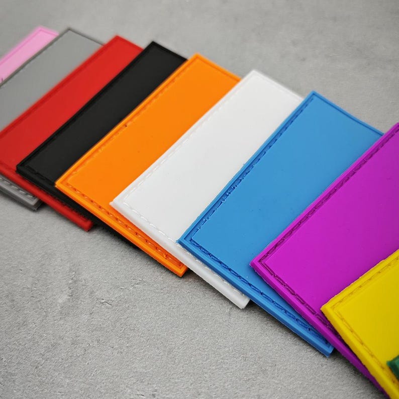 May include: A set of nine blank rectangular patches in various colours, including pink, grey, red, black, orange, white, blue, purple, and yellow. The patches have a stitched border and are arranged in a row, with the pink patch at the top and the yellow patch at the bottom.