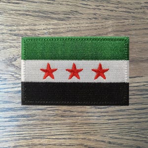 Puede incluir: Un parche bordado verde, blanco y negro con tres estrellas rojas. El parche es rectangular y tiene un borde cosido.