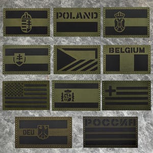 May include: A set of olive green and black embroidered patches displaying national flags and country names. The patches include flags of the USA, Poland, Belgium, and others, with detailed emblems and text.