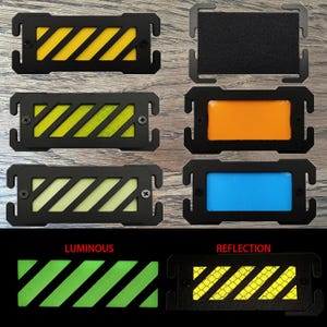 Puede incluir: Seis parches rectangulares negros con insertos de diferentes colores. Los insertos son amarillos, verdes, blancos, naranjas, azules y un patrón de cuadrícula amarillo y negro. Los parches están etiquetados como "LUMINOUS" y "REFLECTION".