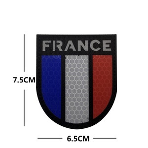 May include: A black and white reflective patch with the word "FRANCE" in white letters and a blue, white, and red French flag design. The patch is 6.5 cm wide and 7.5 cm tall.