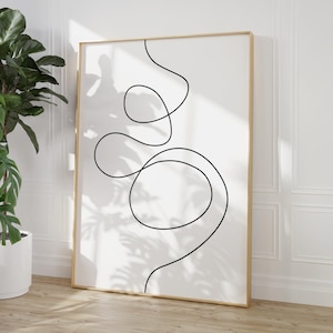 May include: A framed minimalist art print with a continuous black line drawing on a white background. The artwork is in a thin, light-coloured wooden frame. The print is leaning against a white wall, with a potted plant in the foreground.