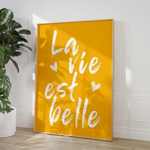 Könnte beinhalten: Ein gerahmter Kunstdruck mit einem leuchtend gelben Hintergrund. Der weiße Text "La vie est belle" wird in handschriftlicher Form mit kleinen Herzillustrationen dargestellt. Der Rahmen hat eine helle Holzfarbe.