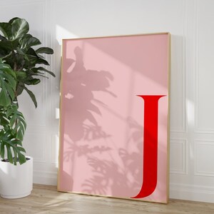 Może przedstawiać: Oprawiony różowy wydruk artystyczny z dużą czerwoną literą "J". Grafika ma złotą ramę i opiera się o białą ścianę. Roślina doniczkowa jest widoczna na pierwszym planie.