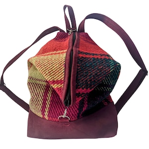 May include: A burgundy and plaid backpack with adjustable straps. The bag features a zippered main compartment and a suede-like bottom. The plaid pattern includes red, yellow, and green hues. The backpack is designed for carrying items.