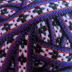 May include: Close-up of a woven fabric with a repeating pattern of black, white, pink, blue, and red. The fabric has a textured, hand-woven look.