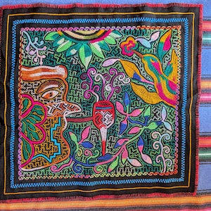 May include: A square, embroidered textile with a black border and a vibrant design. The central image features a colourful hummingbird, flowers, and a stylised human figure. The background is a dark green with intricate stitching.