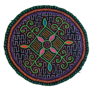 Pode incluir: Um círculo redondo de tecido preto com um design bordado colorido. O design apresenta uma forma de diamante central com quatro motivos florais estilizados em tons de rosa, laranja, verde e azul. O diamante é rodeado por um padrão repetitivo de pequenos quadrados pretos.