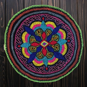 Peut inclure: Un motif circulaire brodé de couleurs vives sur fond noir. Le motif présente un élément central à quatre points, entouré de cercles concentriques de différentes couleurs, notamment rouge, bleu, vert, jaune et rose.