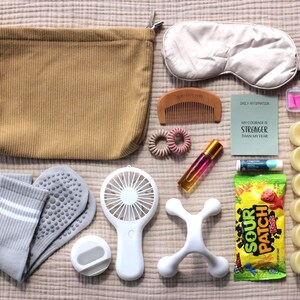 May include: A tan corduroy pouch, a beige sleep mask, a wooden comb, two pink hair coils, a glass roller bottle, a card with the text MY COURAGE IS STRONGER THAN MY FEAR, a small container, gray socks, a white fan, a white massager, lip balm, and candy.