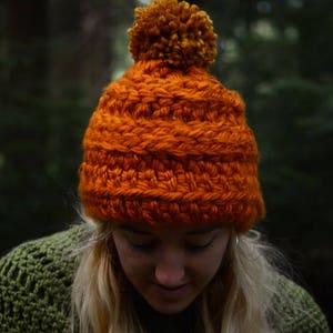 K&ouml;nnte beinhalten: Eine leuchtend orangefarbene H&auml;kelm&uuml;tze mit einem flauschigen orangefarbenen Bommel oben. Die M&uuml;tze ist aus dickem Garn gefertigt und hat ein strukturiertes Stichmuster.