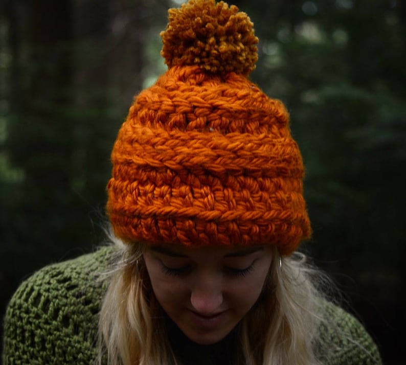 K&ouml;nnte beinhalten: Eine leuchtend orangefarbene H&auml;kelm&uuml;tze mit einem flauschigen orangefarbenen Bommel oben. Die M&uuml;tze ist aus dickem Garn gefertigt und hat ein strukturiertes Stichmuster.