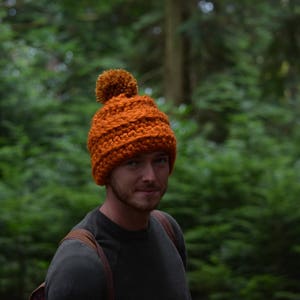 K&ouml;nnte beinhalten: Ein Mann tr&auml;gt eine leuchtend orangefarbene Strickm&uuml;tze mit einem braunen Bommel. Die M&uuml;tze hat ein Zopfmuster. Er tr&auml;gt ein dunkelgraues Hemd und steht in einem Wald.