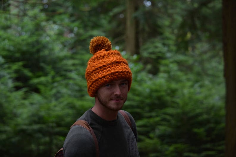 K&ouml;nnte beinhalten: Ein Mann tr&auml;gt eine leuchtend orangefarbene Strickm&uuml;tze mit einem braunen Bommel. Die M&uuml;tze hat ein Zopfmuster. Er tr&auml;gt ein dunkelgraues Hemd und steht in einem Wald.