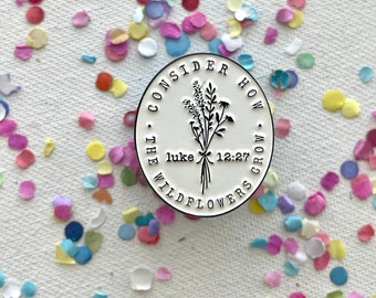 Puede incluir: Un pin de metal blanco con el texto "Considera cómo crecen las flores silvestres - Lucas 12:27" y un dibujo de flores silvestres.