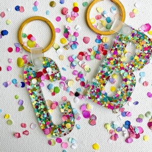 May include: Two gold keychains with clear resin letters that spell "CB". The letters are filled with colorful confetti.