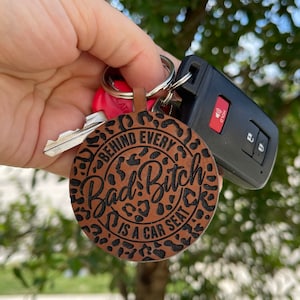 May include: A brown leather keychain with a black leopard print design. The keychain has the text "Behind every Bad Bitch is a car seat".
