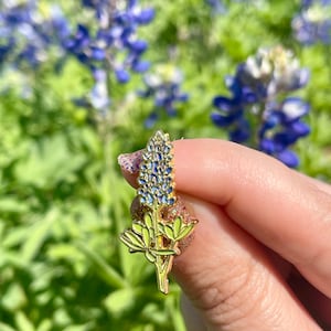 May include: Gold-toned enamel pin featuring a bluebonnet flower with green leaves. The pin is a popular symbol of Texas.