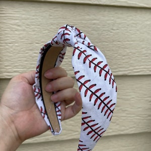 May include: White headband with a red baseball stitch pattern. The headband is tied in a knot at the top.