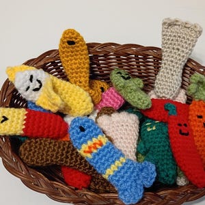 May include: A brown woven basket filled with a collection of small, colorful, crocheted plush toys. The items include various shapes and figures, such as a yellow banana, a blue fish, a red chili pepper, and an orange carrot, all featuring embroidered faces.