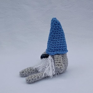 May include: A handmade crochet gnome doll with a blue pointed hat, gray body, and long white beard. The gnome is sitting down with its legs extended. The doll is made of soft yarn and has a whimsical appearance.