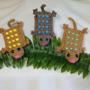 May include: Three crocheted lizard-shaped figures in tan, gray, and light brown. Each figure features a rectangular body adorned with rows of plastic beads in yellow, blue, or green, dark round eyes, and a small pink stitched nose.