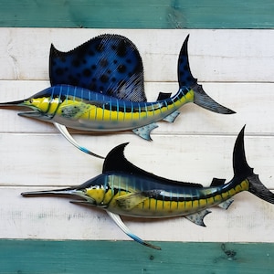 May include: Two blue and yellow sailfish mounted on a white wood background. The sailfish have black spots on their dorsal fins.