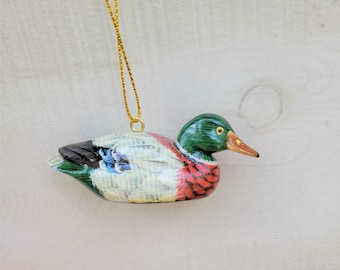 May include: A wooden duck ornament with a green head, red breast, and white body. The duck has a yellow beak and black eyes. The ornament is hanging from a gold string.