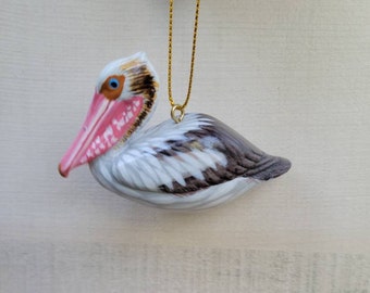 May include: A ceramic pelican ornament with a gold string for hanging. The pelican has a white body with brown and gray accents on the wings and back. The beak is pink, and the eye is blue. The ornament is a decorative item.