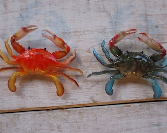May include: Two decorative crab figurines. One is bright orange with yellow legs and claws, the other is dark green with blue claws. Both have their claws raised, as if in a playful gesture. The crabs are placed on a light-colored, weathered wooden surface.