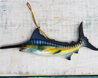 May include: A blue and yellow marlin fish ornament with a gold string for hanging. The fish has a long, pointed beak and a dorsal fin.