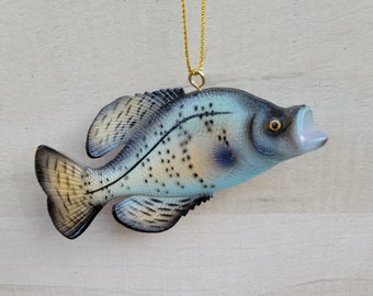 May include: A blue and black plastic fish ornament with a gold string for hanging. The fish has a speckled pattern and a slightly open mouth.