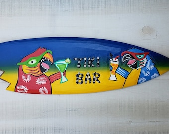 May include: A wooden surfboard painted with a blue, yellow, and green gradient. Two parrots are painted on the surfboard, each holding a tropical drink. The text "TIKI BAR" is painted in yellow letters on the surfboard.
