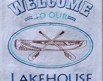 May include: Embroidered kitchen towel with the text "Welcome to our Lakehouse" in blue and brown thread. A canoe with crossed paddles is embroidered in the center of the towel.